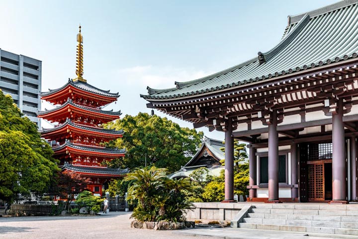 A view of Tochoji Temple in Fukuoka, Japan, featuring a five-story red pagoda and a traditional Japanese temple building with a grey tiled roof.