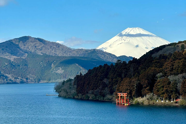 This image captures the stunning landscape of Hakone, Japan, with a clear view of Mount Fuji's majestic peak. In the foreground, Lake Ashi's blue waters meet a shoreline featuring a vibrant red torii gate.