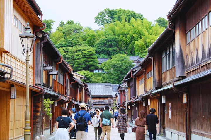 A street scene in Kanazawa's Higashi Chaya District, featuring traditional wooden buildings, a lantern, and people strolling along the narrow street.