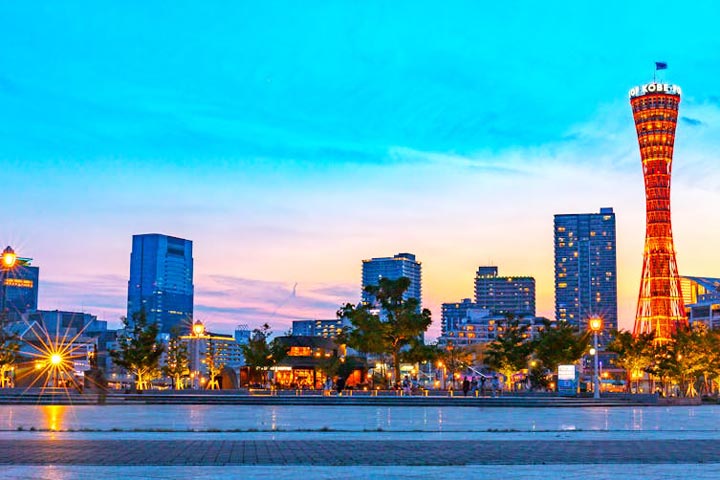 The illuminated Kobe Port Tower stands prominently in the evening skyline of Kobe, Japan, with city lights reflecting on the waterfront under a colorful sky.