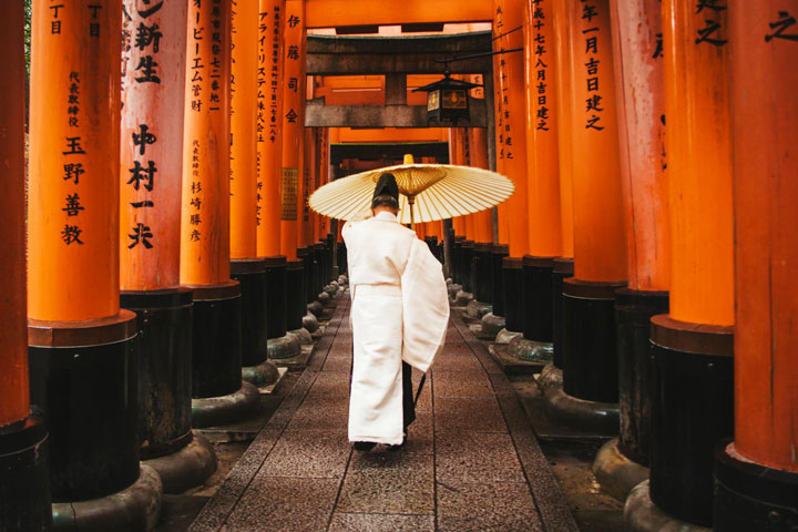 A person in white walks through the vibrant orange torii gates of Fushimi Inari Shrine in Kyoto, Japan.