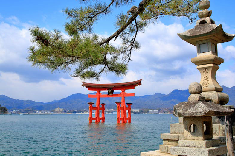 The iconic orange torii gate of Itsukushima Shrine rises from the water near Miyajima Island in Hiroshima, Japan, with a stone lantern and a branch visible in the foreground.