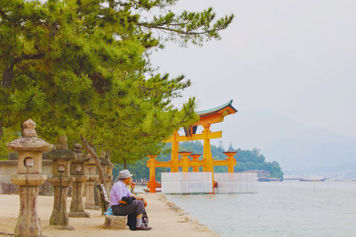 A person sits on a bench near stone lanterns, looking out at the iconic orange floating torii gate of Itsukushima Shrine in Hiroshima, Japan.