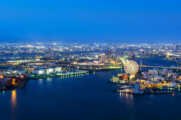 A vibrant view of Osaka, Japan, at night, with the city lights reflecting on the water and a Ferris wheel prominently featured.
