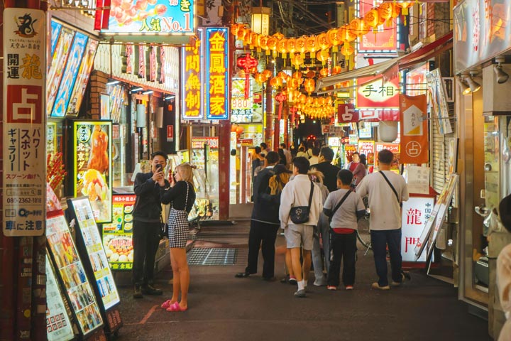 A bustling street scene at night in Yokohama, Japan, filled with colorful signs, lanterns, and people walking along the narrow alleyway.